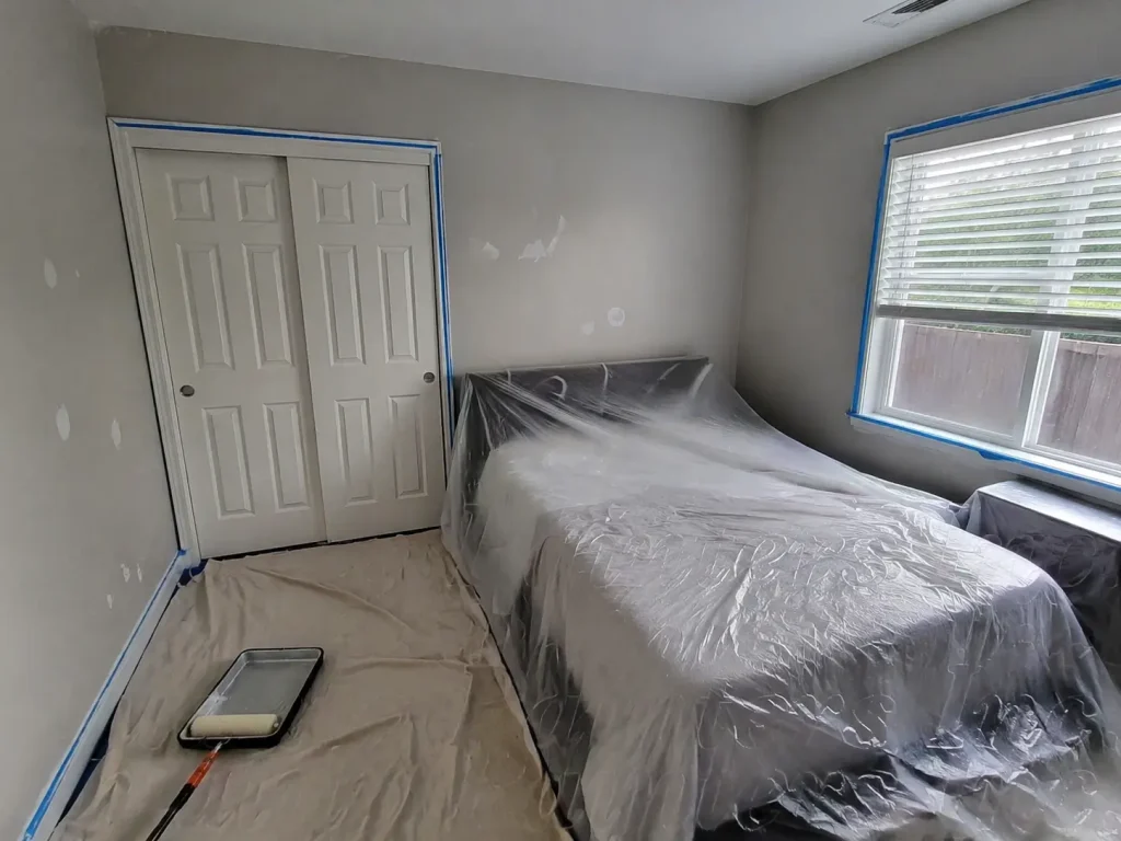 Bedroom prepared for interior painting with covered furniture, taped trim, patched walls, and drop cloths.