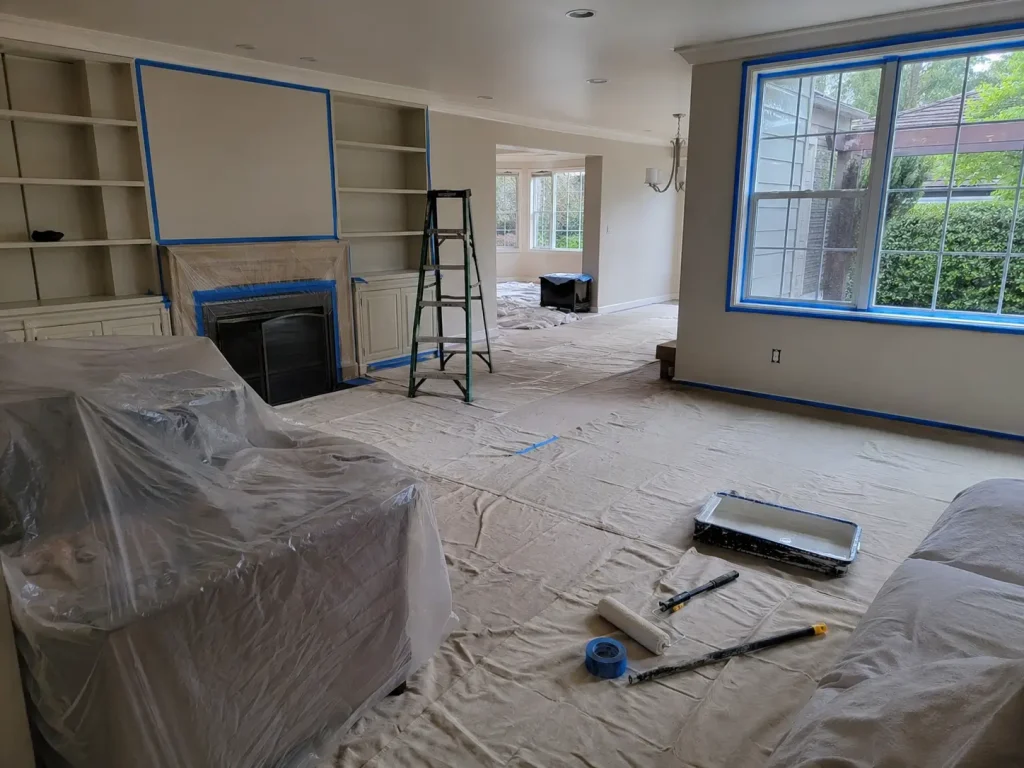 Living room prepared for painting with covered furniture, taped windows, built-ins, ladder, rollers, and floor protection.