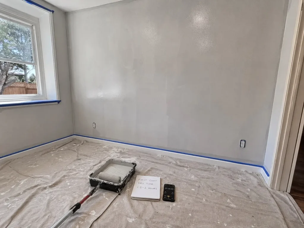 Interior wall painting setup with a roller tray, taped baseboards, drop cloths, and a wall drying before the second coat.