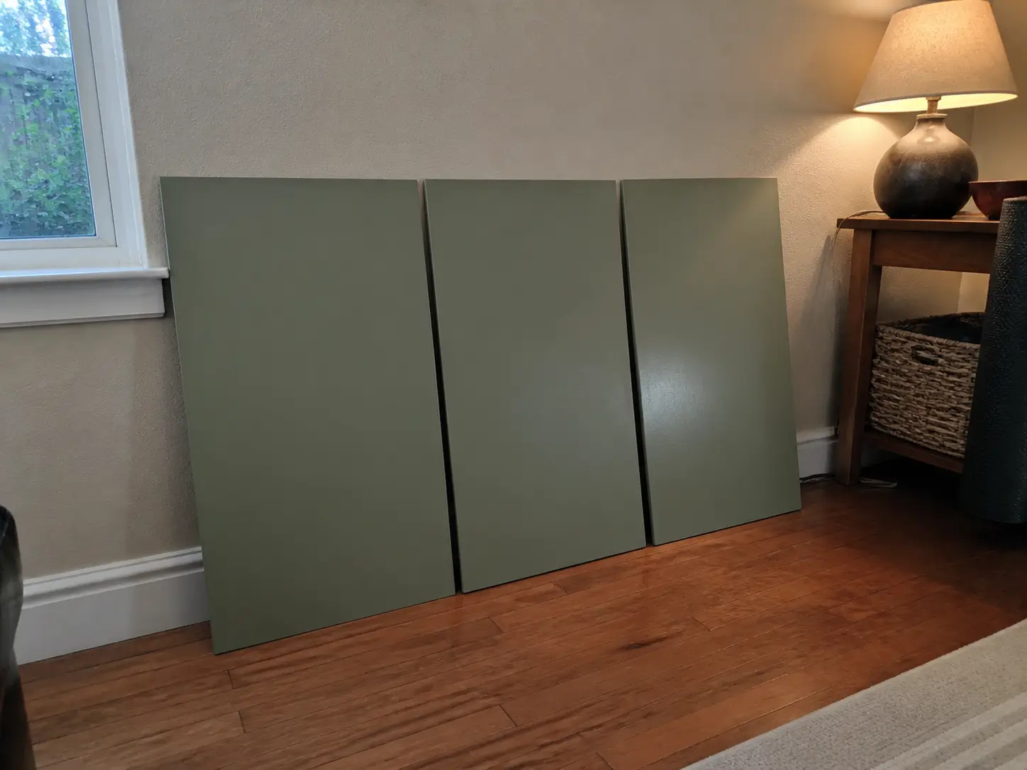 Three sage-green paint sample boards in different sheens under natural window light and a table lamp.