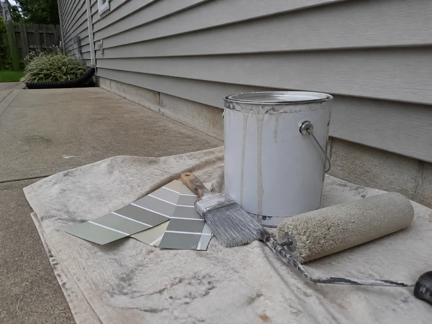 Exterior acrylic paint, roller, brush, and neutral color swatches arranged beside vinyl siding.