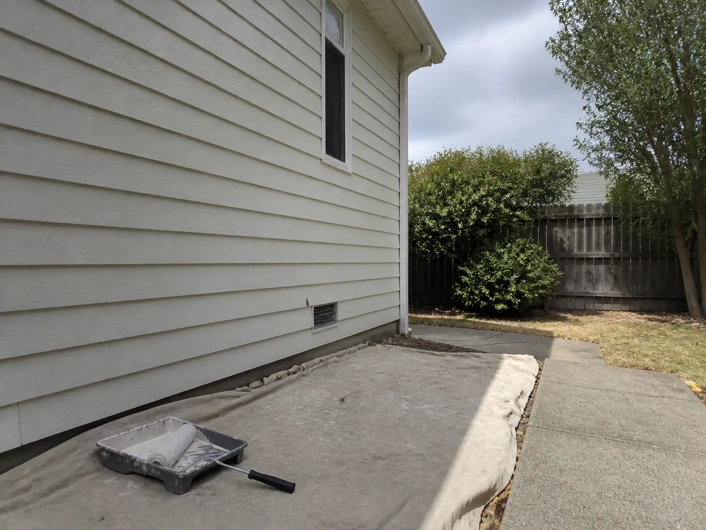 Paint tray and roller set in the shade beside dry vinyl siding on a mild day.