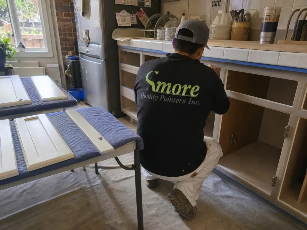 Amore Painters contractor prepares and paints kitchen cabinet frames while removed cabinet doors rest on padded work tables.